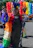 A person dressed in vibrant, colorful attire with an elaborate feathered headpiece stands waving during a festive parade. The outfit features a rainbow-colored cape and various sparkling accessories. In the background, large clusters of balloons in rainbow colors enhance the celebratory atmosphere.