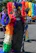 A person dressed in vibrant, colorful attire with an elaborate feathered headpiece stands waving during a festive parade. The outfit features a rainbow-colored cape and various sparkling accessories. In the background, large clusters of balloons in rainbow colors enhance the celebratory atmosphere.