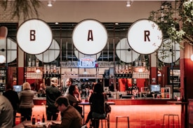 A lively bar scene with a warm, inviting atmosphere. Large circular light fixtures with the letters B, A, and R hang prominently above the counter. Several patrons are gathered around the bar, conversing and enjoying drinks. The bar itself is well-stocked with various liquors and glasses, and there are plants adding a touch of greenery to the space.