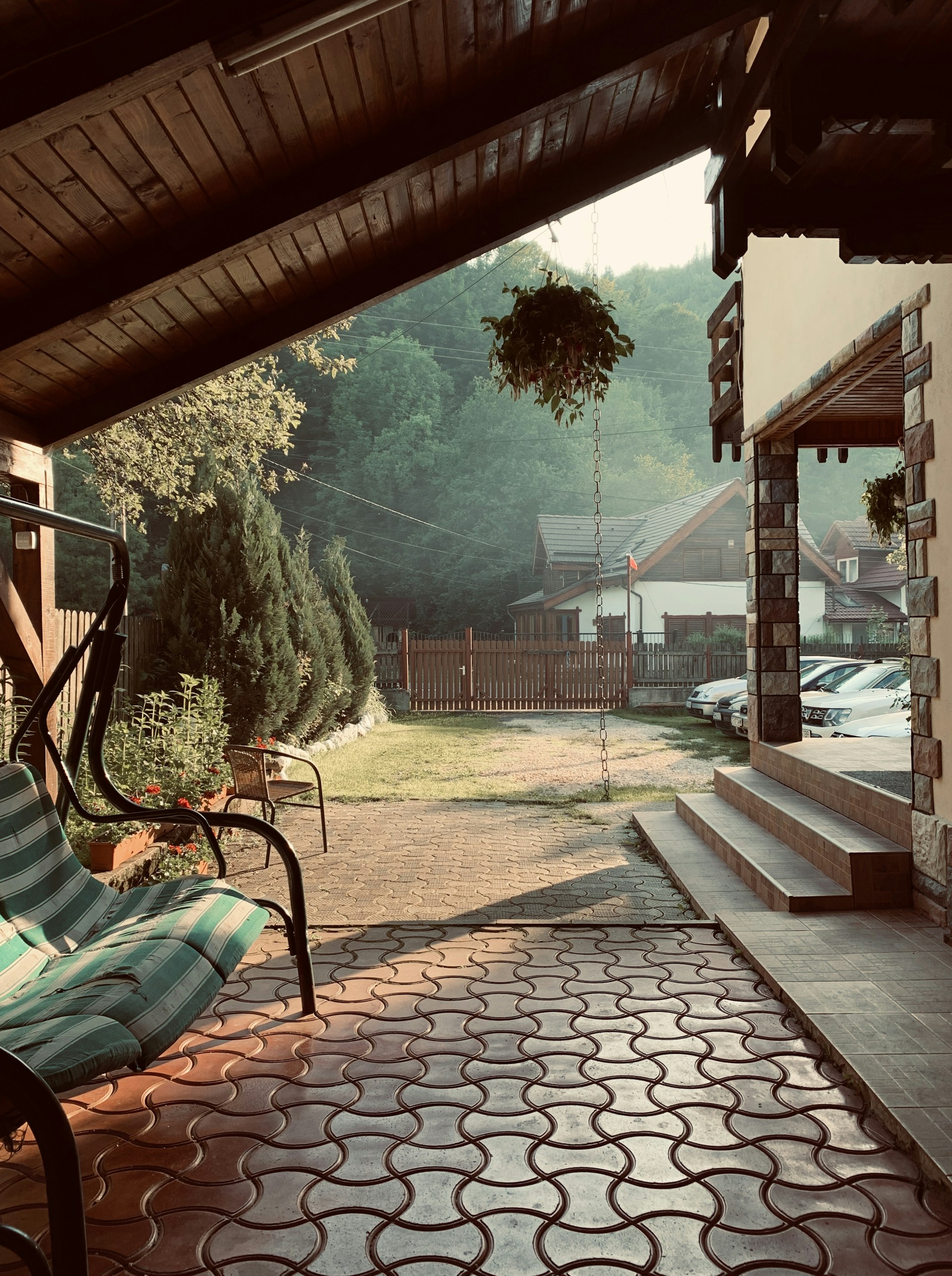 A cozy outdoor area with a wooden ceiling above a patio featuring patterned tiles. A green and white striped swing chair is to the left, complemented by a small garden with flowers and shrubs along the edges. Beyond the patio, there's a grassy yard leading to a fence, with several parked cars visible to the right. The scene is serene, with gentle sunlight filtering through, creating a warm and relaxing atmosphere.
