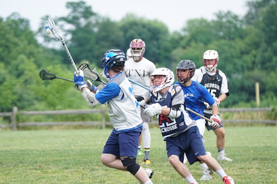 A group of young men are playing a game of lacrosse on a green field. One player in a white and blue jersey is in possession of the ball, while others are in pursuit. They are wearing helmets, gloves, and protective gear. The players appear focused and in motion, with trees and a wooden fence in the background.