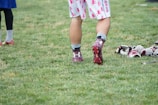 A person wearing sports shoes and patterned shorts stands on a grassy field, with various sports equipment like gloves and a helmet placed on the ground nearby.