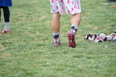 A person wearing sports shoes and patterned shorts stands on a grassy field, with various sports equipment like gloves and a helmet placed on the ground nearby.