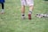 A person wearing sports shoes and patterned shorts stands on a grassy field, with various sports equipment like gloves and a helmet placed on the ground nearby.