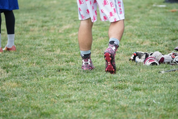 A person wearing sports shoes and patterned shorts stands on a grassy field, with various sports equipment like gloves and a helmet placed on the ground nearby.
