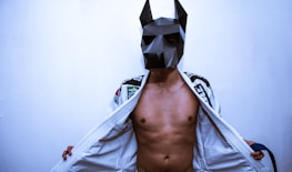A person wearing a geometric, angular mask reminiscent of an animal figure. The individual is dressed in a martial arts gi, which is open to reveal their bare torso. The background is a plain, light-colored wall.