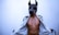A person wearing a geometric, angular mask reminiscent of an animal figure. The individual is dressed in a martial arts gi, which is open to reveal their bare torso. The background is a plain, light-colored wall.