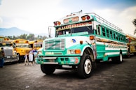 A vibrantly painted bus with turquoise and white colors is prominently displayed, featuring decorative elements and intricate patterns. In the background, several other similarly styled buses are parked, creating a lively scene. A few people, including children, are present near the buses, suggesting a busy, communal atmosphere.