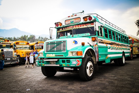 A vibrantly painted bus with turquoise and white colors is prominently displayed, featuring decorative elements and intricate patterns. In the background, several other similarly styled buses are parked, creating a lively scene. A few people, including children, are present near the buses, suggesting a busy, communal atmosphere.