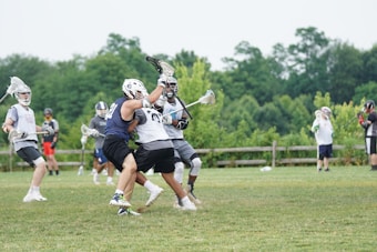 A group of young athletes playing lacrosse on a grassy field, surrounded by lush green trees. In the foreground, two players engage closely, each holding a lacrosse stick, while others in the background prepare to join the action or observe. They wear protective gear including helmets and padded jerseys.