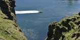 A yellow speedboat travels swiftly across a large body of water, leaving a white wake behind. On either side of the waterway, steep, grassy cliffs rise, covered in lush green vegetation. The water reflects sunlight, creating sparkles on its surface.