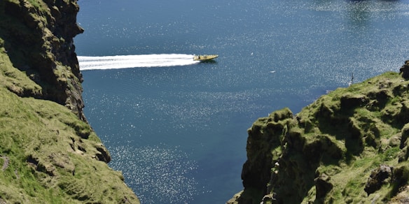 A yellow speedboat travels swiftly across a large body of water, leaving a white wake behind. On either side of the waterway, steep, grassy cliffs rise, covered in lush green vegetation. The water reflects sunlight, creating sparkles on its surface.