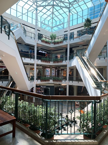 A multi-level shopping mall interior with a glass ceiling, escalators, and potted plants decorating the walkways on each level. Various shops with prominent signs are visible around the open center space.