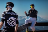 Two people are dressed in sporty attire with reflective goggles, set against a scenic backdrop of snowy mountains. One person is standing with their back to the viewer, wearing a shirt with 'DragBlack' on it, while the other is sitting casually on a wooden railing, engaged in conversation. The scene captures the essence of outdoor adventure and leisure.
