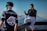 Two people are dressed in sporty attire with reflective goggles, set against a scenic backdrop of snowy mountains. One person is standing with their back to the viewer, wearing a shirt with 'DragBlack' on it, while the other is sitting casually on a wooden railing, engaged in conversation. The scene captures the essence of outdoor adventure and leisure.