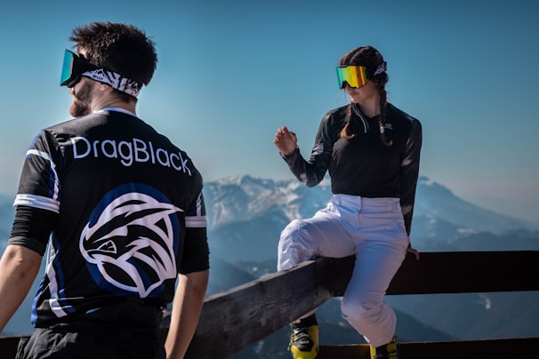 Two people are dressed in sporty attire with reflective goggles, set against a scenic backdrop of snowy mountains. One person is standing with their back to the viewer, wearing a shirt with 'DragBlack' on it, while the other is sitting casually on a wooden railing, engaged in conversation. The scene captures the essence of outdoor adventure and leisure.