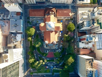 An aerial view of a city block featuring a prominent, historic building with a large central dome and red-tiled roof. The surrounding area includes high-rise buildings, streets lined with cars, and patches of greenery, possibly small parks or gardens. The layout creates a contrast between urban development and natural elements, with streets forming a structured grid.