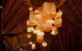 A warm, ambient setting with multiple woven, rattan-style pendant lights hanging from a high wooden ceiling. The lights vary in size and shape, providing a cozy and inviting atmosphere.