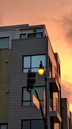 A modern multi-story building with large windows is shown at sunset. The sky is a vivid orange, creating a striking backdrop for the building. In front of the building, a traffic light is visible, displaying both green and yellow lights. An adjacent street lamp emits a warm glow.