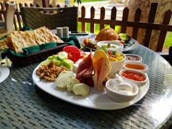 A table is set with a variety of breakfast items on a white plate including slices of cucumber, tomato, walnuts, cheese, and an assortment of deli meats. Accompanying these are several small bowls filled with various dips and jams. On the table, there are also flatbreads in a basket and a croissant on a separate plate. The outdoor setting gives a pleasant and relaxed dining ambiance.