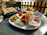 A table is set with a variety of breakfast items on a white plate including slices of cucumber, tomato, walnuts, cheese, and an assortment of deli meats. Accompanying these are several small bowls filled with various dips and jams. On the table, there are also flatbreads in a basket and a croissant on a separate plate. The outdoor setting gives a pleasant and relaxed dining ambiance.