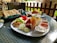 A table is set with a variety of breakfast items on a white plate including slices of cucumber, tomato, walnuts, cheese, and an assortment of deli meats. Accompanying these are several small bowls filled with various dips and jams. On the table, there are also flatbreads in a basket and a croissant on a separate plate. The outdoor setting gives a pleasant and relaxed dining ambiance.