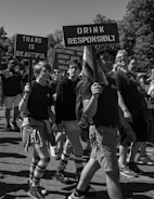 A group of people participating in a march holding signs with messages promoting responsibility and LGBTQ+ pride. The signs read 'Trans is Beautiful' and 'Drink Responsibly'. Participants are wearing casual attire, some with striped socks and shorts, walking along a sunny street. Trees can be seen in the background.