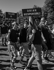 A group of people participating in a march holding signs with messages promoting responsibility and LGBTQ+ pride. The signs read 'Trans is Beautiful' and 'Drink Responsibly'. Participants are wearing casual attire, some with striped socks and shorts, walking along a sunny street. Trees can be seen in the background.