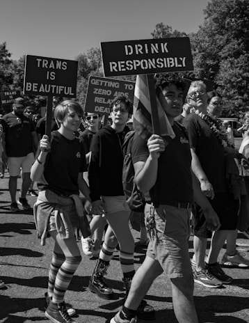 A group of people participating in a march holding signs with messages promoting responsibility and LGBTQ+ pride. The signs read 'Trans is Beautiful' and 'Drink Responsibly'. Participants are wearing casual attire, some with striped socks and shorts, walking along a sunny street. Trees can be seen in the background.