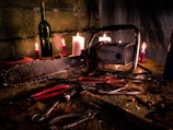 A chainsaw is placed on a cluttered wooden table, surrounded by various tools like pliers and scissors. The background features a stone wall and several lit candles that cast a dim, eerie light. A wine bottle is positioned among the candles, contributing to the scene's haunting atmosphere.