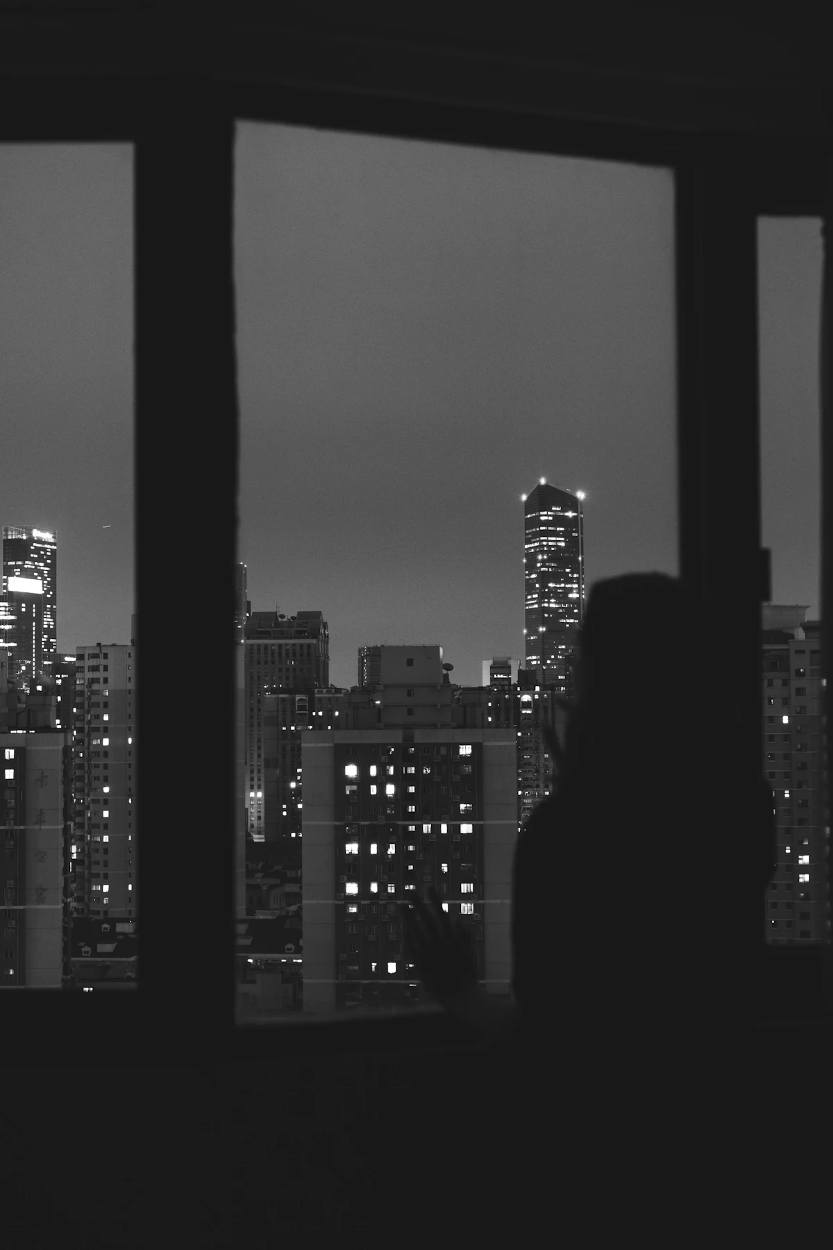 A person standing at a dark window looking out over a city full of lit apartment windows at night
