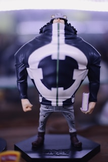 A detailed figurine of a character with a large upper body, wearing a distinct outfit with a black and white design and a hat with ear-like features. The figure holds a small book in one hand and stands on a base labeled with 'One Piece.' The focus is on the intricate costume detailing and painted features.