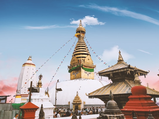 A grand stupa with ornate golden decorations and multiple tiers stands prominently, surrounded by smaller shrines. Colorful prayer flags string across the sky, adding vibrancy against the clear blue backdrop. The architecture features intricate details, typical of Buddhist shrines, with a blend of red, white, and gold elements. Numerous visitors move about in the area, suggesting a lively atmosphere.