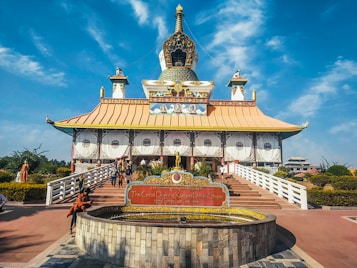 The image showcases a large ornate stupa with a golden roof and intricate decorative elements, surrounded by a landscaped garden. People are walking along a staircase leading up to the entrance, and a sign with text in multiple languages is prominently displayed at the front.