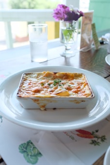 A creamy, golden-brown baked dish served in a rectangular ceramic ramekin resting on a white plate, placed on a wooden table. Next to it, there's a clear glass of water and a small vase with purple and white flowers in the background.