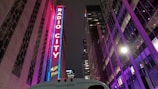 A tall, illuminated sign with the words 'Radio City Music Hall' stands vertically on a building. The surroundings feature modern architecture with bright, colorful lights reflecting on glass surfaces. The scene is set at night, and the atmosphere is vibrant with purple and pink neon lighting.