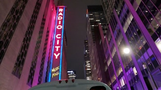 A tall, illuminated sign with the words 'Radio City Music Hall' stands vertically on a building. The surroundings feature modern architecture with bright, colorful lights reflecting on glass surfaces. The scene is set at night, and the atmosphere is vibrant with purple and pink neon lighting.
