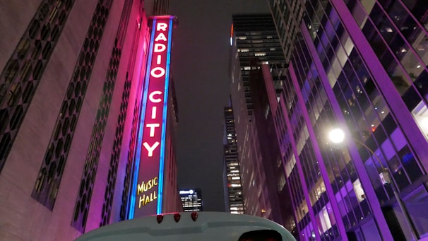 A tall, illuminated sign with the words 'Radio City Music Hall' stands vertically on a building. The surroundings feature modern architecture with bright, colorful lights reflecting on glass surfaces. The scene is set at night, and the atmosphere is vibrant with purple and pink neon lighting.