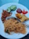 A white plate features a serving of thin rice noodles, garnished with leafy greens. To the side, there are two crispy fried appetizers. Additional accompaniments include sliced cucumber, roasted peanuts with small dried fish, a dollop of red chili sauce, and a blob of ketchup. A metal fork is placed on the plate.