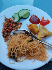 A white plate features a serving of thin rice noodles, garnished with leafy greens. To the side, there are two crispy fried appetizers. Additional accompaniments include sliced cucumber, roasted peanuts with small dried fish, a dollop of red chili sauce, and a blob of ketchup. A metal fork is placed on the plate.