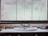 A vintage-style bathroom sink is set against a marble countertop, featuring ornate silver faucets. In the background, a large window with sheer curtains allows diffused natural light to enter. Various toiletries, including a soap dispenser and a toothbrush holder, are neatly arranged on the countertop.