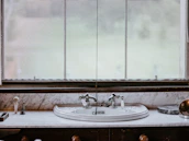A vintage-style bathroom sink is set against a marble countertop, featuring ornate silver faucets. In the background, a large window with sheer curtains allows diffused natural light to enter. Various toiletries, including a soap dispenser and a toothbrush holder, are neatly arranged on the countertop.