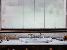 A vintage-style bathroom sink is set against a marble countertop, featuring ornate silver faucets. In the background, a large window with sheer curtains allows diffused natural light to enter. Various toiletries, including a soap dispenser and a toothbrush holder, are neatly arranged on the countertop.
