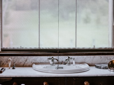 A vintage-style bathroom sink is set against a marble countertop, featuring ornate silver faucets. In the background, a large window with sheer curtains allows diffused natural light to enter. Various toiletries, including a soap dispenser and a toothbrush holder, are neatly arranged on the countertop.