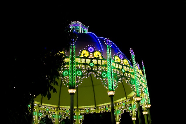 A vibrant, illuminated pavilion structure featuring intricate designs in green and blue lights against a dark night background. The canopy is adorned with floral patterns and pointed spires, creating a festive and lively atmosphere.