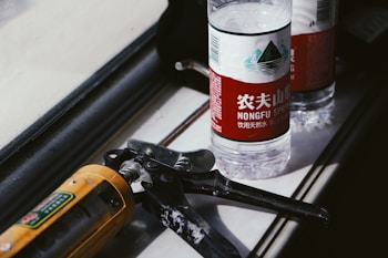A bottle of Nongfu Spring water with a red label sits on a surface next to a caulking gun. The scene is lit by natural light, casting distinct shadows and highlighting the textures of the objects.