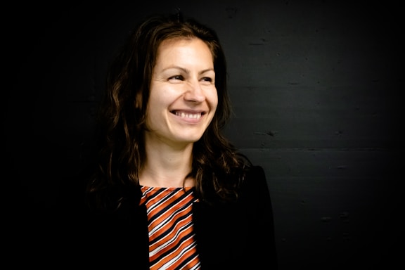 A woman with long, wavy brown hair is smiling and looking slightly to her left. She is wearing a black blazer over a multicolored striped top with shades of orange, white, and black. The background is a dark shade, contrasting with her lighter skin tone.