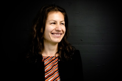 A woman with long, wavy brown hair is smiling and looking slightly to her left. She is wearing a black blazer over a multicolored striped top with shades of orange, white, and black. The background is a dark shade, contrasting with her lighter skin tone.