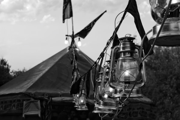 A series of vintage lanterns hang from a string in front of a large tent. Flags and lights are also visible, suggesting an outdoor setting with a rustic or traditional theme. The scene appears to be in black and white, enhancing a nostalgic or historical feel.