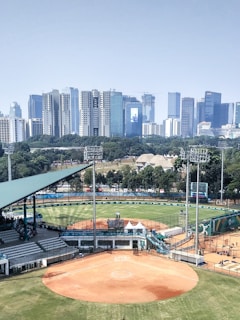 A baseball or softball stadium is in the foreground with a well-maintained field and seating areas. Beyond the stadium, there is a cityscape featuring numerous high-rise buildings and skyscrapers, providing a contrast between the sports facility and the urban environment. Trees and greenery surround the stadium, adding a natural element to the scene.